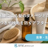 明るい窓辺の木のテーブルに、ヘアオイルの瓶と木製ブラシ、麦わら帽子が並ぶ静物イメージ